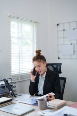 Siyah takım elbiseli bir iş kadını, masasında bilgisayarla çalışırken telefonla konuşuyor. Profesyonel ofis ortamında birden fazla görev ve verimliliği somutlaştırıyor..