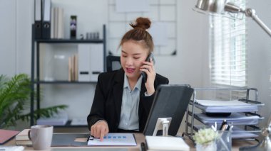 Siyah takım elbiseli bir iş kadını, masasında bilgisayarla çalışırken telefonla konuşuyor. Profesyonel ofis ortamında birden fazla görev ve verimliliği somutlaştırıyor..