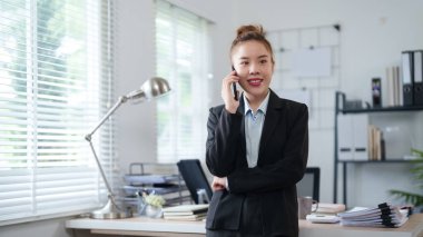 Siyah takım elbiseli bir iş kadını masasının yanında duruyor, telefonda konuşuyor, modern ofis ortamında güven ve profesyonellik yayıyor..