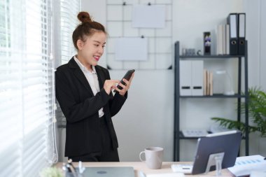 Siyah takım elbiseli bir iş kadını pencerenin yanında duruyor ve modern ofis ortamında akıllı telefonunu kontrol ediyor.