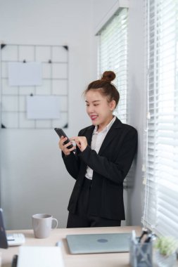 Siyah takım elbiseli bir iş kadını pencerenin yanında duruyor ve modern ofis ortamında akıllı telefonunu kontrol ediyor.