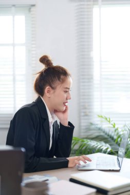 Siyah takım elbiseli bir iş kadını masasında çalışıyor. Kararlı bir göreve odaklanıyor. Etrafı ofis malzemeleri ve belgelerle çevrili.