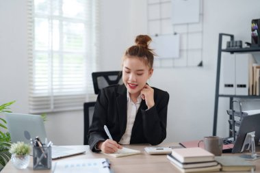 Siyah takım elbiseli kendine güvenen bir iş kadını masasında oturmuş, çevrimiçi bir toplantıya katılırken gülümsüyor ve modern bir ofiste masanın üzerinde notlar ve belgeler düzenleniyor..