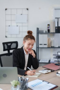 Siyah takım elbiseli kendine güvenen bir iş kadını masasında oturmuş, çevrimiçi bir toplantıya katılırken gülümsüyor ve modern bir ofiste masanın üzerinde notlar ve belgeler düzenleniyor..