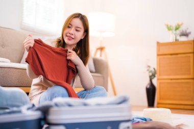 Genç bir kadın yerde oturuyor, valizini düzenlerken paketleme listesindeki eşyaları kontrol ediyor, her şeyin yolunda olduğundan emin oluyor..