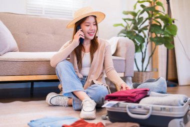 Şapka takan genç bir kadın yerde oturuyor, telefonda konuşurken gülümsüyor, bavulunu toplarken, rahat bir oturma odasında yolculuğa hazırlanıyor..