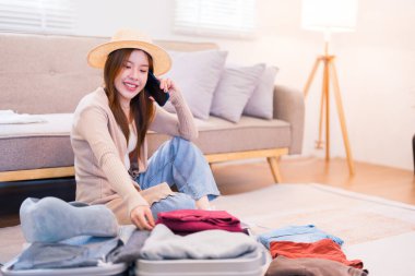 Şapka takan genç bir kadın yerde oturuyor, telefonda konuşurken gülümsüyor, bavulunu toplarken, rahat bir oturma odasında yolculuğa hazırlanıyor..