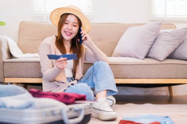 Genç bir kadın yerde oturuyor, telefonda konuşuyor ve biniş kartına bakıyor, yanında bavuluyla bir yolculuğa hazırlanırken gülümsüyor..