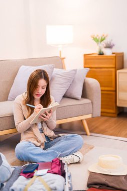 Genç bir kadın yerde oturuyor, valizini düzenlerken paketleme listesindeki eşyaları kontrol ediyor, her şeyin yolunda olduğundan emin oluyor..