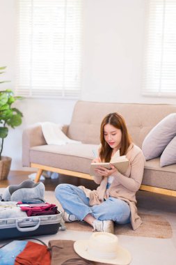 Genç bir kadın yerde oturuyor, valizini düzenlerken paketleme listesindeki eşyaları kontrol ediyor, her şeyin yolunda olduğundan emin oluyor..