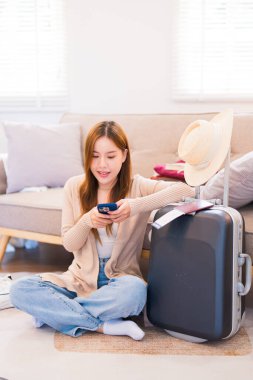 Genç bir kadın valizinin yanında oturuyor, akıllı telefonunu kullanarak seyahat bilgilerini arıyor, yaklaşan seyahatine hazır..