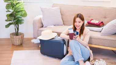 Genç bir kadın yerde rahat bir şekilde oturuyor, elinde bir pasaport ve akıllı telefonunu kullanarak seyahat bilgilerini arıyor, yaklaşan yolculuğuna hazırlanıyor..