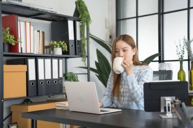 Asyalı bir kadın iş kadını, bilgisayarını açıp masasında otururken fincandan yudumlayarak modern ofis ortamında bir anlık rahatlama keyfi yapıyor.