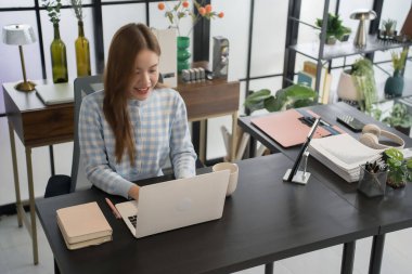 Mavi kareli gömlekli Asyalı bir kadın işkadını, düzenli dosyalar ve ofis malzemeleriyle çevrili modern bir ofis masasında dizüstü bilgisayarıyla çalışmaya odaklanıyor..