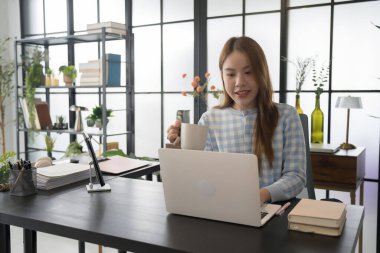 Asyalı bir kadın iş kadını, bilgisayarını açıp masasında otururken fincandan yudumlayarak modern ofis ortamında bir anlık rahatlama keyfi yapıyor.