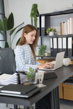 Mavi kareli gömlekli Asyalı bir kadın işkadını, düzenli dosyalar ve ofis malzemeleriyle çevrili modern bir ofis masasında dizüstü bilgisayarıyla çalışmaya odaklanıyor..