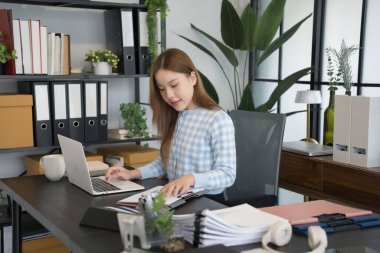 Mavi kareli gömlekli Asyalı bir kadın işkadını masasında bilgisayarını açıp, modern ofis ortamındaki çalışmalarına konsantre olarak evrakları inceliyor.