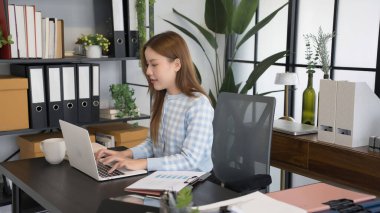 Mavi kareli gömlekli Asyalı bir kadın işkadını, düzenli dosyalar ve ofis malzemeleriyle çevrili modern bir ofis masasında dizüstü bilgisayarıyla çalışmaya odaklanıyor..