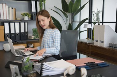 Mavi kareli gömlekli Asyalı bir kadın işkadını masasında bilgisayarını açıp, modern ofis ortamındaki çalışmalarına konsantre olarak evrakları inceliyor.