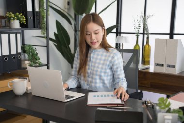 Mavi kareli gömlekli Asyalı bir kadın işkadını masasında bilgisayarını açıp, modern ofis ortamındaki çalışmalarına konsantre olarak evrakları inceliyor.
