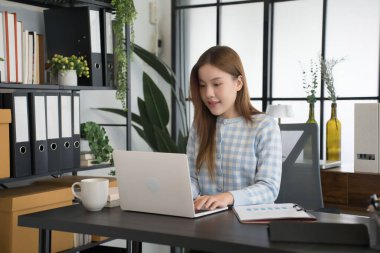 Mavi kareli gömlekli Asyalı bir kadın işkadını, düzenli dosyalar ve ofis malzemeleriyle çevrili modern bir ofis masasında dizüstü bilgisayarıyla çalışmaya odaklanıyor..