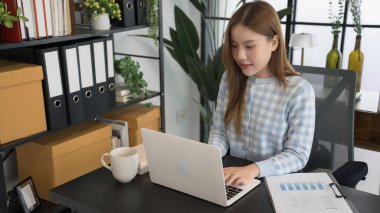 Mavi kareli gömlekli Asyalı bir kadın işkadını, düzenli dosyalar ve ofis malzemeleriyle çevrili modern bir ofis masasında dizüstü bilgisayarıyla çalışmaya odaklanıyor..