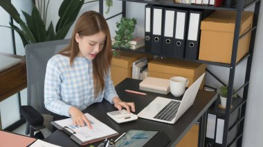 Asyalı bir kadın iş kadını masasında oturuyor ve birden fazla iş üzerinde çalışıyor. Belgelerine, hesap makinesine ve dizüstü bilgisayarına odaklanmış, modern ve düzenli bir çalışma alanına dalmış..