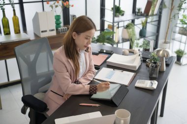 Asyalı bir kadın işkadını masasında oturmuş, kağıtlar ve dizüstü bilgisayarla çevriliyken bir tablete notlar yazıyor. Ofis ayarları çağdaş ve iyi düzenlenmiş.