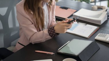 Asyalı bir iş kadını, masasında çalışırken bilgisayarla, belgelerle ve kırtasiyeyle çevrili akıllı telefonunu kontrol eder..