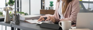 Asyalı bir iş kadını, modern bir ofisteki masasında dijital tablete yazıyor. Belgeler ve ofis malzemeleriyle işine odaklanmış durumda. 