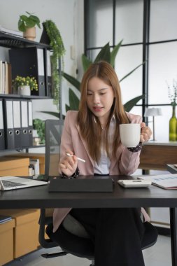 Neşeli bir Asyalı iş kadını masasında çalışırken, parlak, modern bir ofiste bir fincan kahvenin tadını çıkarıyor. O, yüzünde bir gülümseme ile işleriyle meşgul.