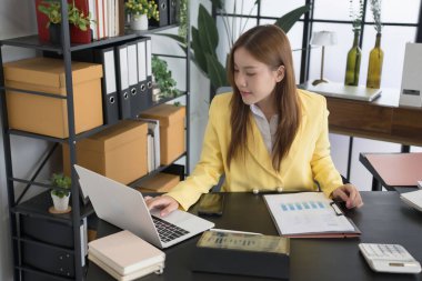 Sarı ceket giyen Asyalı bir iş kadını masasında çalışıyor. Dosyaları ve bilgisayarındaki verileri analiz ediyor. Masası çeşitli belgeler, bir hesap makinesi ve bir kahve fincanı ile düzenlenmiş.,