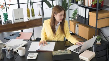 Sarı ceket giyen Asyalı bir iş kadını masasında çalışıyor. Dosyaları ve bilgisayarındaki verileri analiz ediyor. Masası çeşitli belgelerle düzenlenmiş, bir hesap makinesi.,