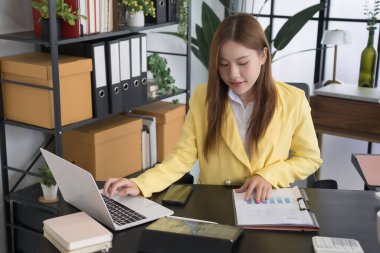 Sarı ceketli Asyalı bir iş kadını masasında organize ofis malzemeleriyle çevrili belgeleri inceliyor. O, işine çok bağlıdır.,