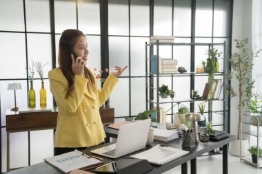 Ofisinde hem akıllı telefonunu hem de laptopunu kullanan sarı ceketli Asyalı bir iş kadını. Masası, modern bir şirket ortamında yoğun ve meşgul olduğunu gösteren belgelerle dolu..