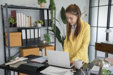 Sarı ceket giyen bir iş kadını masasında dizüstü bilgisayarına odaklanmış duruyor. Çalışma alanı kağıtlar, hesap makinesi ve kahve fincanıyla düzenli.