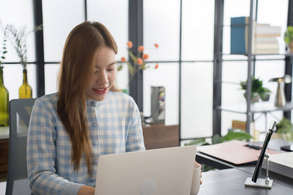 Азиатская деловая женщина в голубой клетчатой рубашке сосредоточена на работе над своим ноутбуком за современным офисным столом в окружении аккуратно организованных файлов и канцелярских принадлежностей.