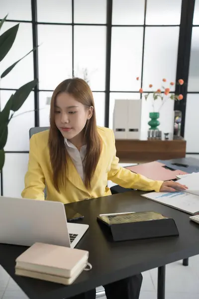 stock image An Asian businesswoman wearing a yellow blazer works at her desk, analyzing charts and data on her laptop. Her desk is organized with various documents, a calculator, and a coffee cup,