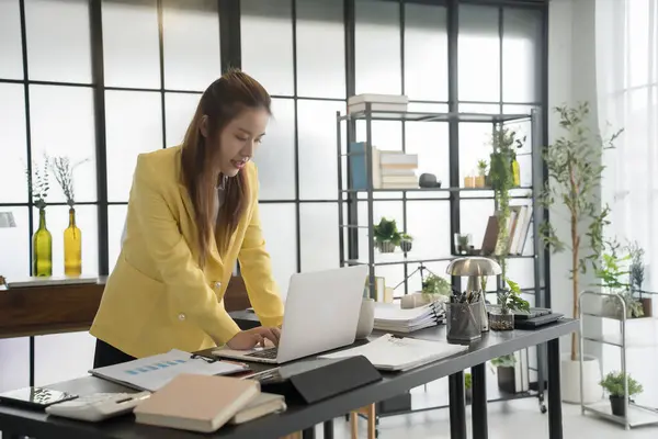 stock image A businesswoman wearing a yellow blazer is standing at her desk, focused on her laptop. Her workspace is neatly organized with papers, a calculator, and a coffee cup