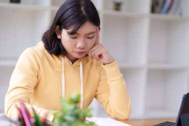 Sarı kapüşonlu genç bir kadın bir masada oturuyor, bir deftere yazıyor. Odaklanmış ve derin düşüncelere dalmış görünüyor. Etrafı temiz ve düzenli bir iç mekanla çevrili..