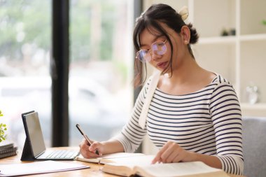 Gözlüklü ve çizgili tişörtlü genç bir kadın bir masada oturuyor, bir deftere yazıyor. Etrafında düzenli bir iç mekan ve arka planda raflar ile odaklanmış görünüyor..