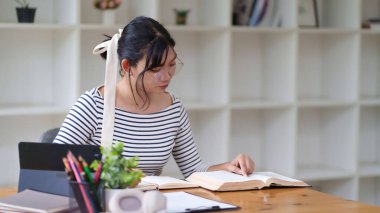 Gözlüklü ve çizgili tişörtlü genç bir kadın bir masada oturuyor, bir deftere yazıyor. Etrafında düzenli bir iç mekan ve arka planda raflar ile odaklanmış görünüyor..