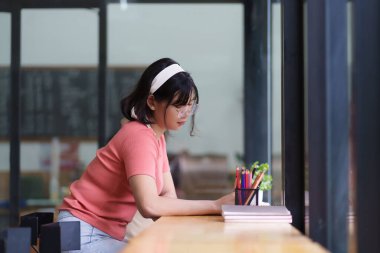 Pembe gömlekli ve gözlüklü genç bir kadın pencerenin yanında oturuyor, bir deftere yazıyor. Oda, sakin bir atmosferle hafifçe aydınlatılıyor ve o işine odaklanıyor.