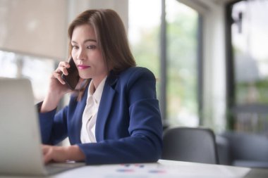Asyalı iş kadınları iş arkadaşlarıyla konuşmak için akıllı telefon kullanıyor ve iş verilerini araştırmak için klavye bilgisayarında yazı yazarken ofis dışındaki yeni girişimlerin yatırım ve pazar stoklarını analiz ediyorlar..
