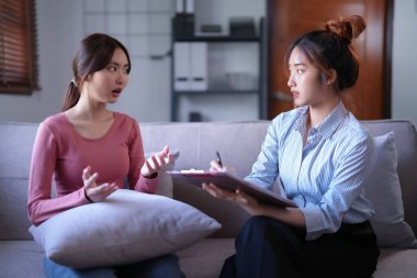 Stresli Asyalı hasta kadınlar doktorlara psikolojik sağlık sorunlarını açıklarken kadın psikoloğlar not panosuna veri notları ve zihinsel sağlık terapisi hakkında danışmanlık alıyorlar..