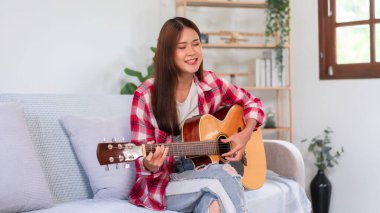 Hobi konsepti, genç Asyalı kadın akustik gitar çalıyor oturma odasındaki kanepede şarkı söylüyor..