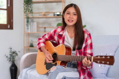 Hobi konsepti, genç Asyalı kadın akustik gitar çalıyor oturma odasındaki kanepede şarkı söylüyor..