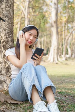 Genç Asyalı kadınlar kulaklık takıyor ve akıllı telefonlarını mutlulukla müzik dinlemek için tutarken parkta bisiklet üstüne bisiklet sürerek seyahat ederken çimenlerin üzerinde rahatlamak için oturuyorlar..