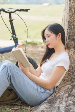 Genç Asyalı kadınlar çime oturup, ağaçlara yaslanarak keyif dolu bir kitap okurken doğa parkında bisiklet üstüne bisiklet sürerek seyahatlerde rahatlıyorlar..