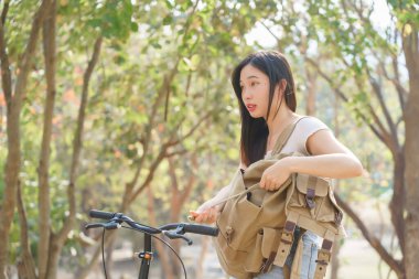 Genç Asyalı kadınlar bisiklete biniyor ve bisiklet hazırlamak için sırtlarında çantayla doğa parkına gidiyorlar. Mutluluk ve rahatlık içinde seyahat ediyorlar. Sağlıklı bir yaşam tarzı için..